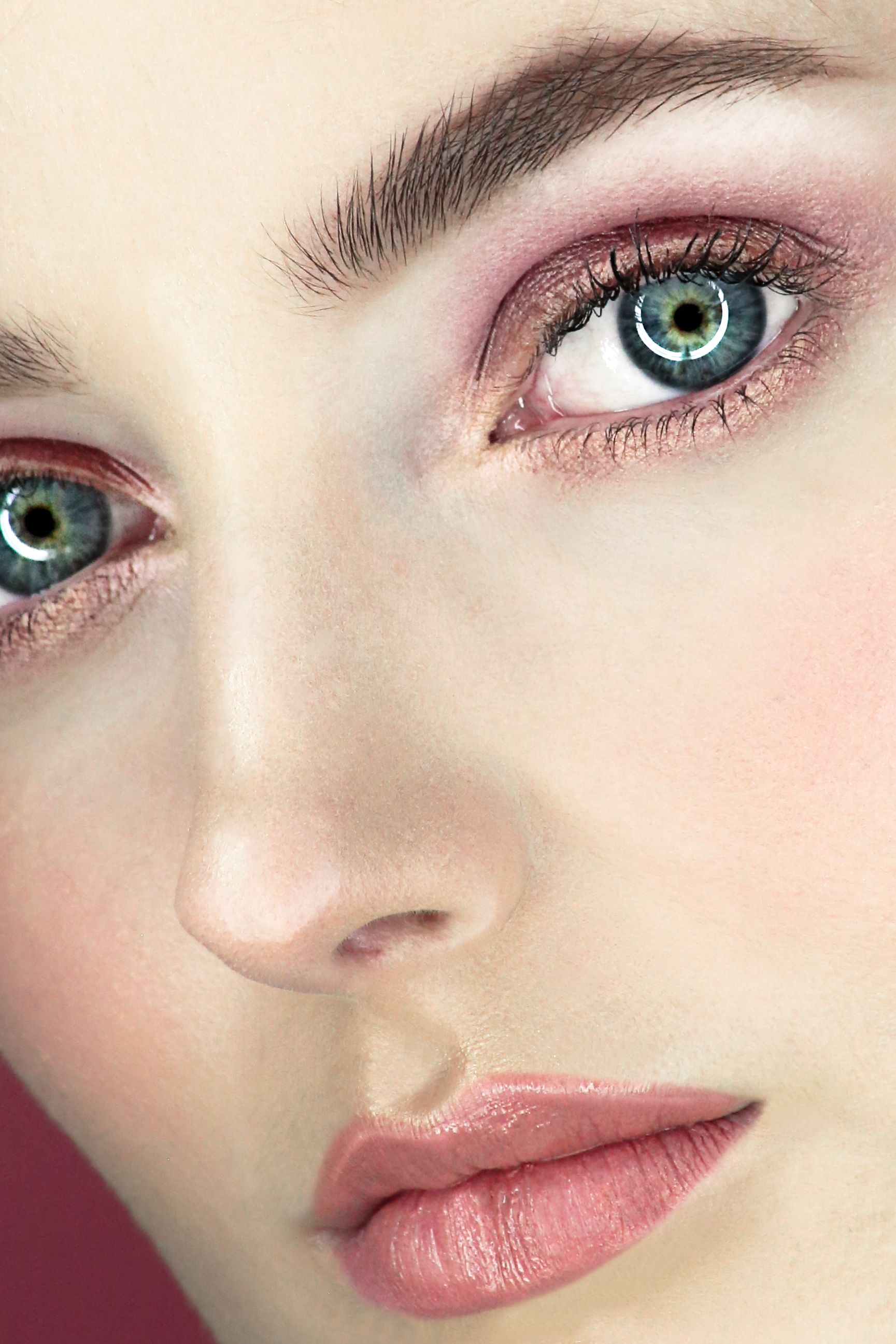 Primer plano de un rostro con ojos azul verdosos, maquillaje rosa suave y labios brillantes.