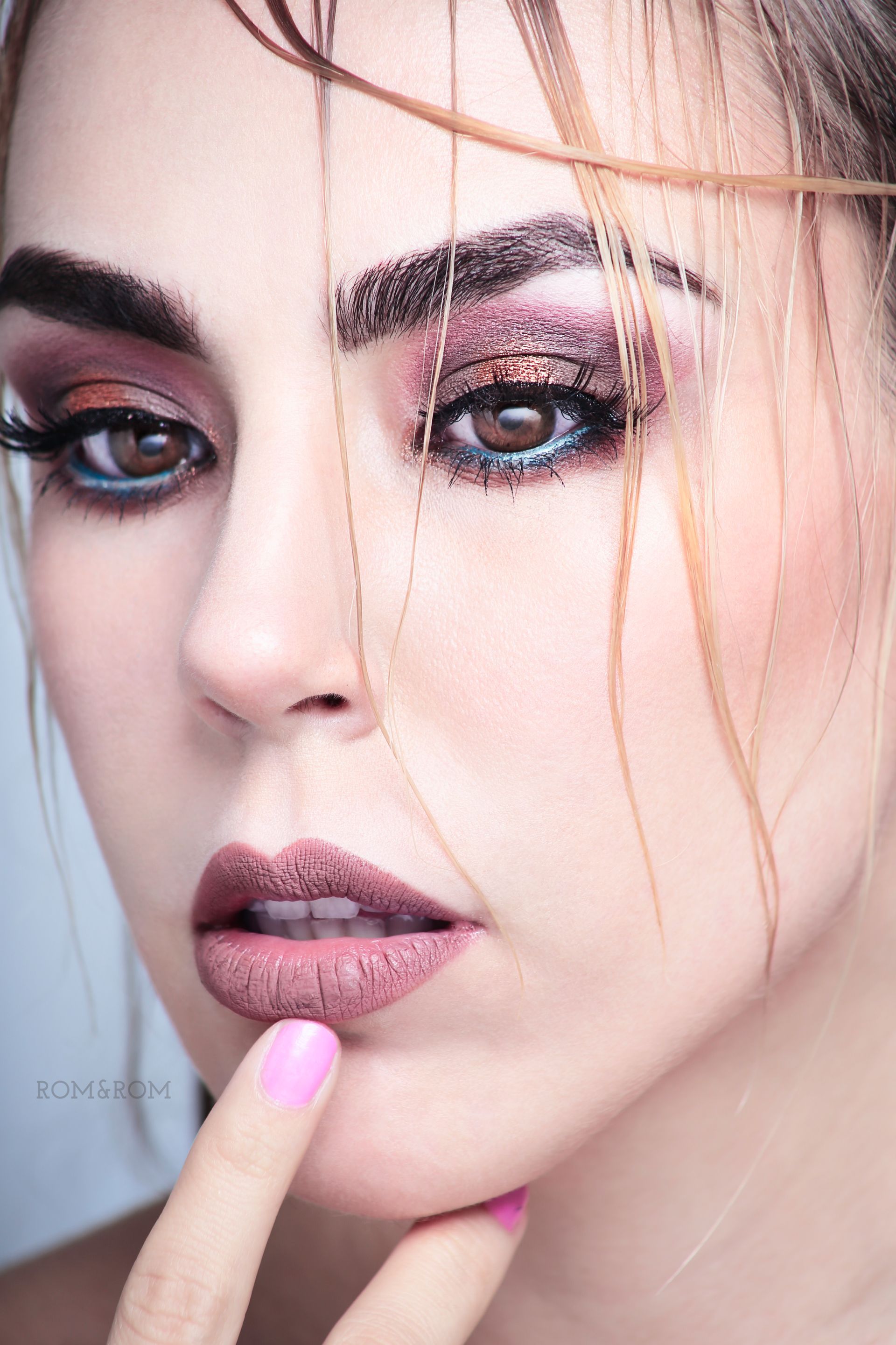 Primer plano de una mujer con un maquillaje de ojos llamativo, mirando a la cámara, con un dedo tocándose el labio inferior.