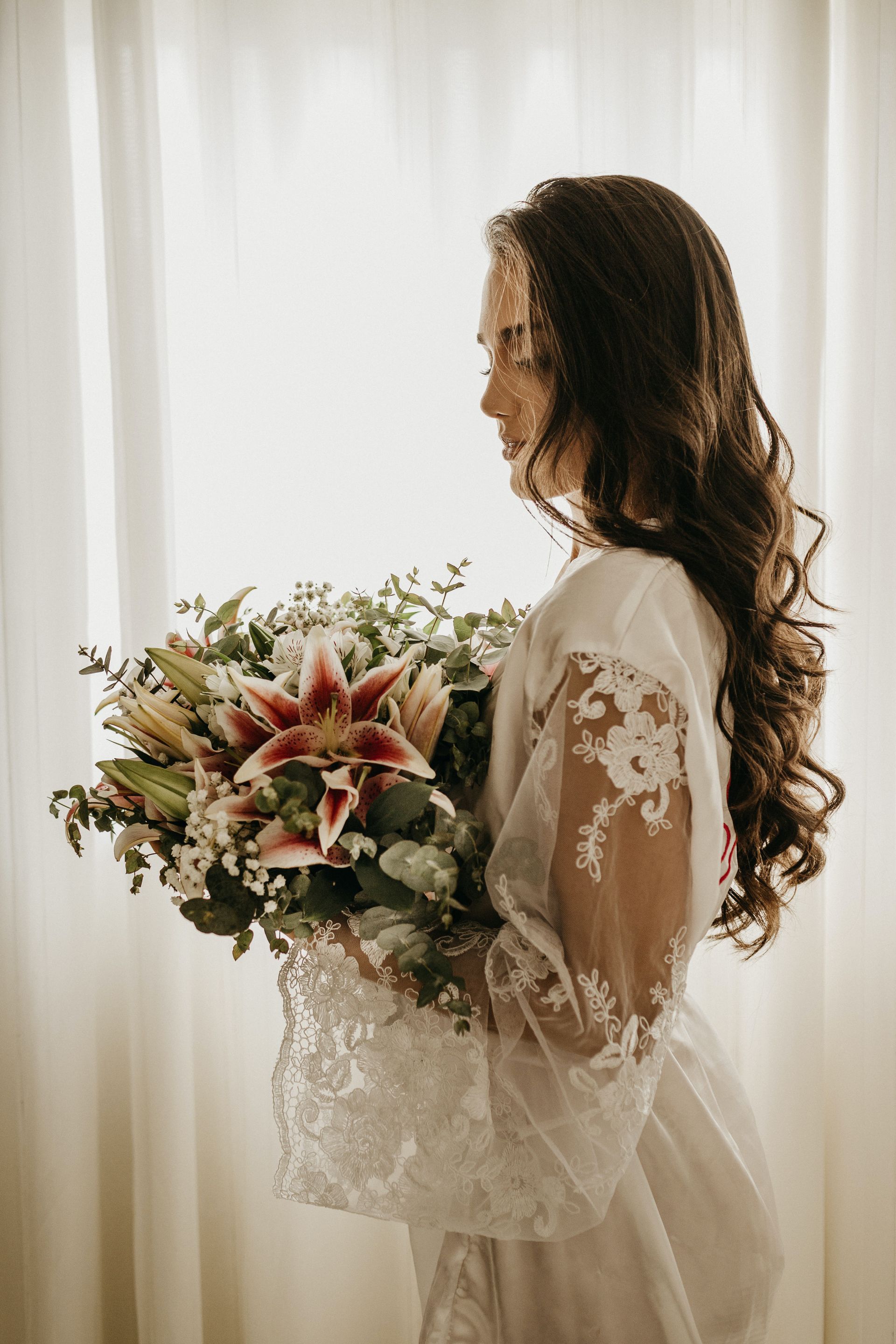 Novia con vestido de encaje sosteniendo un ramo rosa y verde junto a una ventana luminosa.