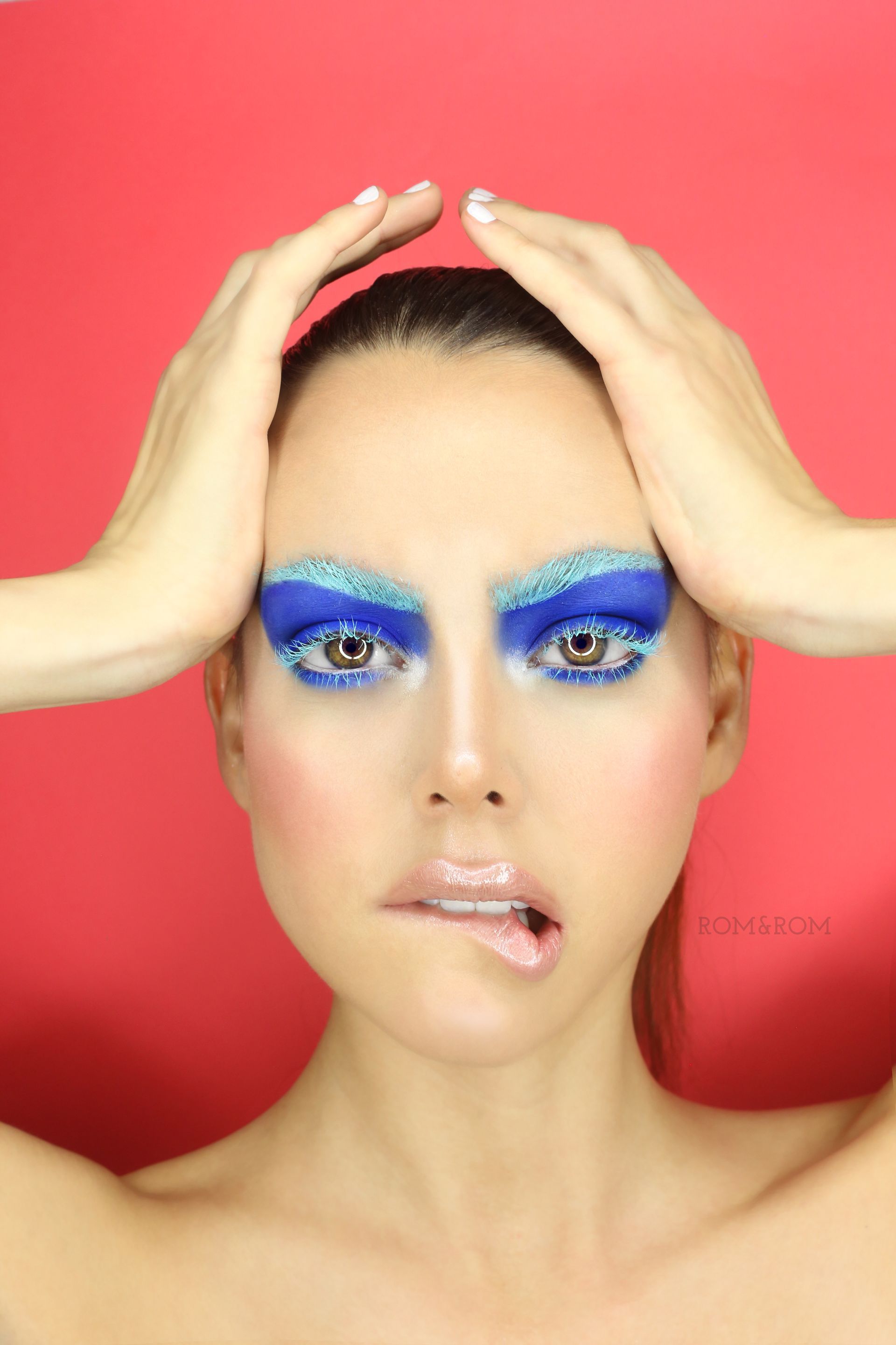 Mujer con maquillaje de ojos azul y labios brillantes, con las manos enmarcando su cabeza sobre un fondo rojo.