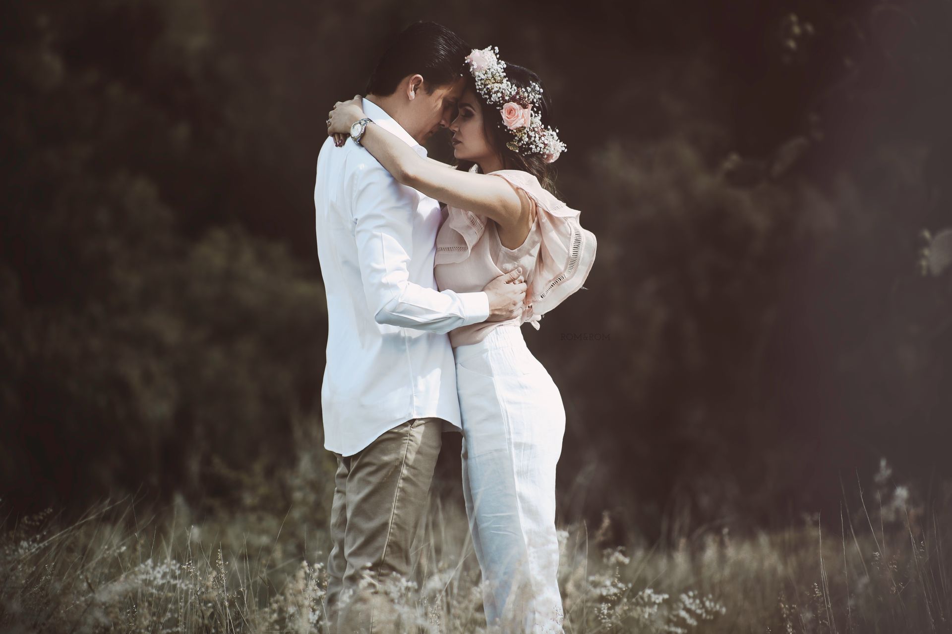 Pareja abrazándose en un campo, vestidos de blanco, con un fondo oscuro difuminado.
