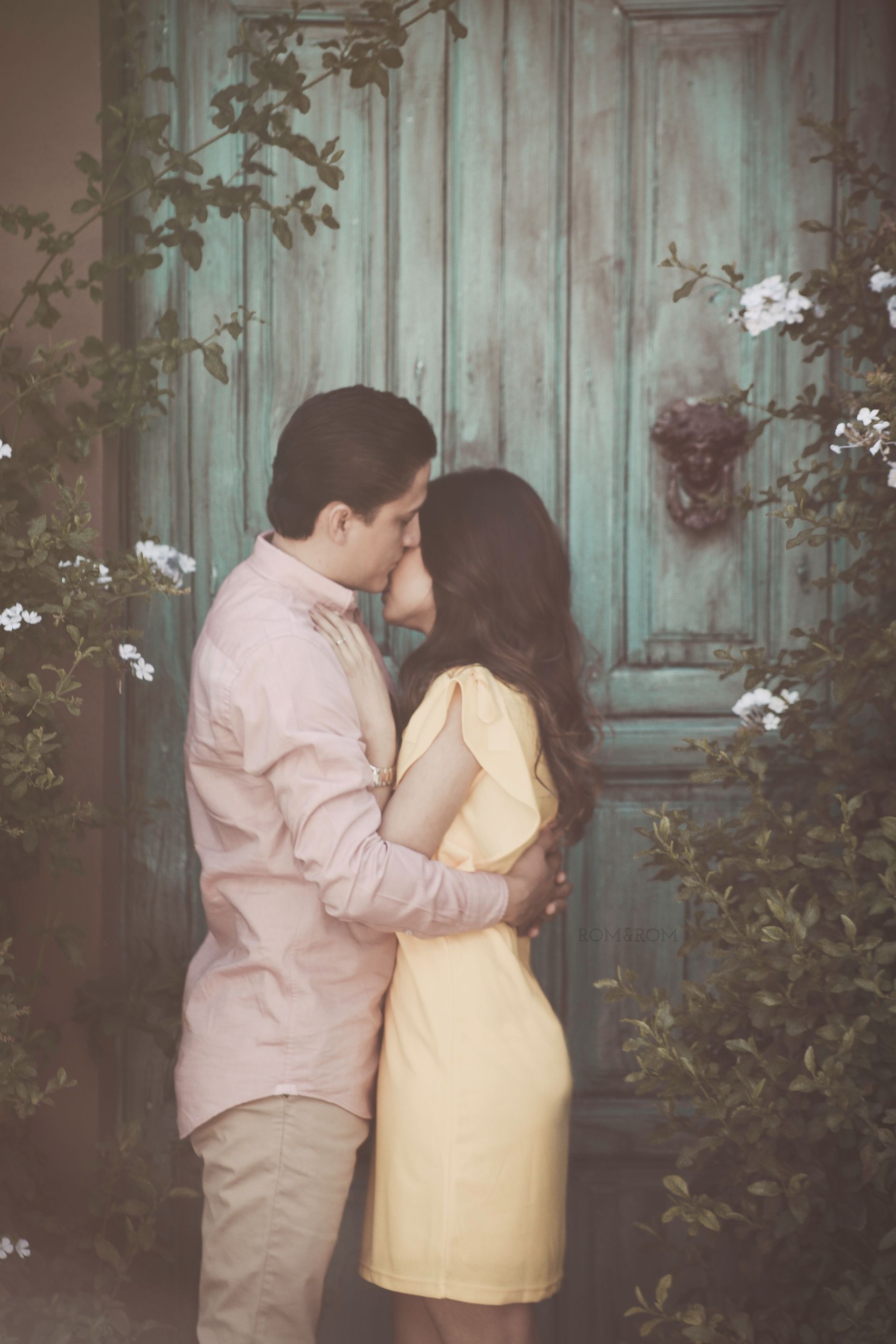 Una pareja se abraza y se besa frente a una puerta turquesa desgastada por el tiempo, rodeada de hiedra y flores.