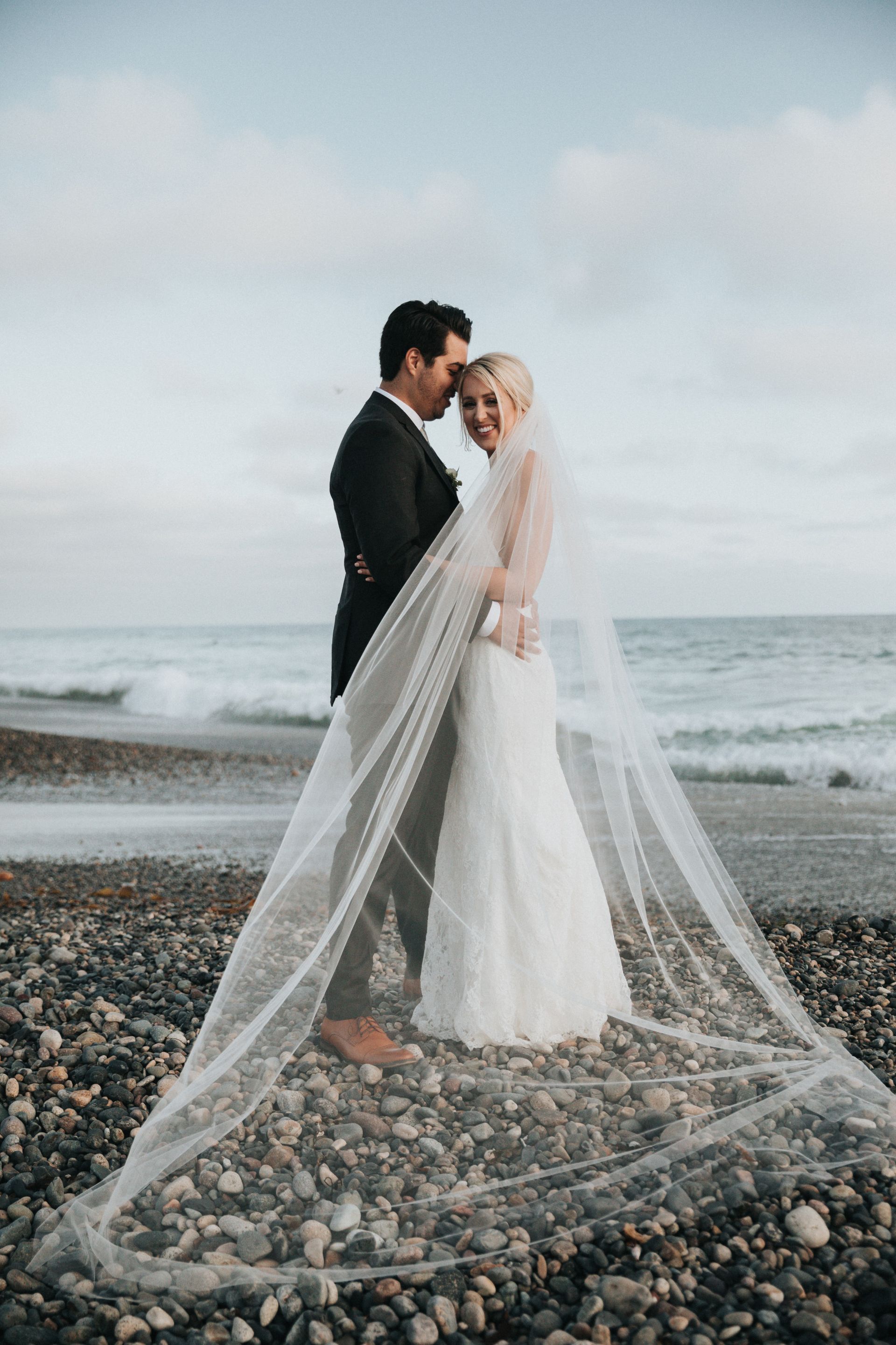 Los novios se abrazan en una playa rocosa, con un largo velo que ondea hacia la orilla.