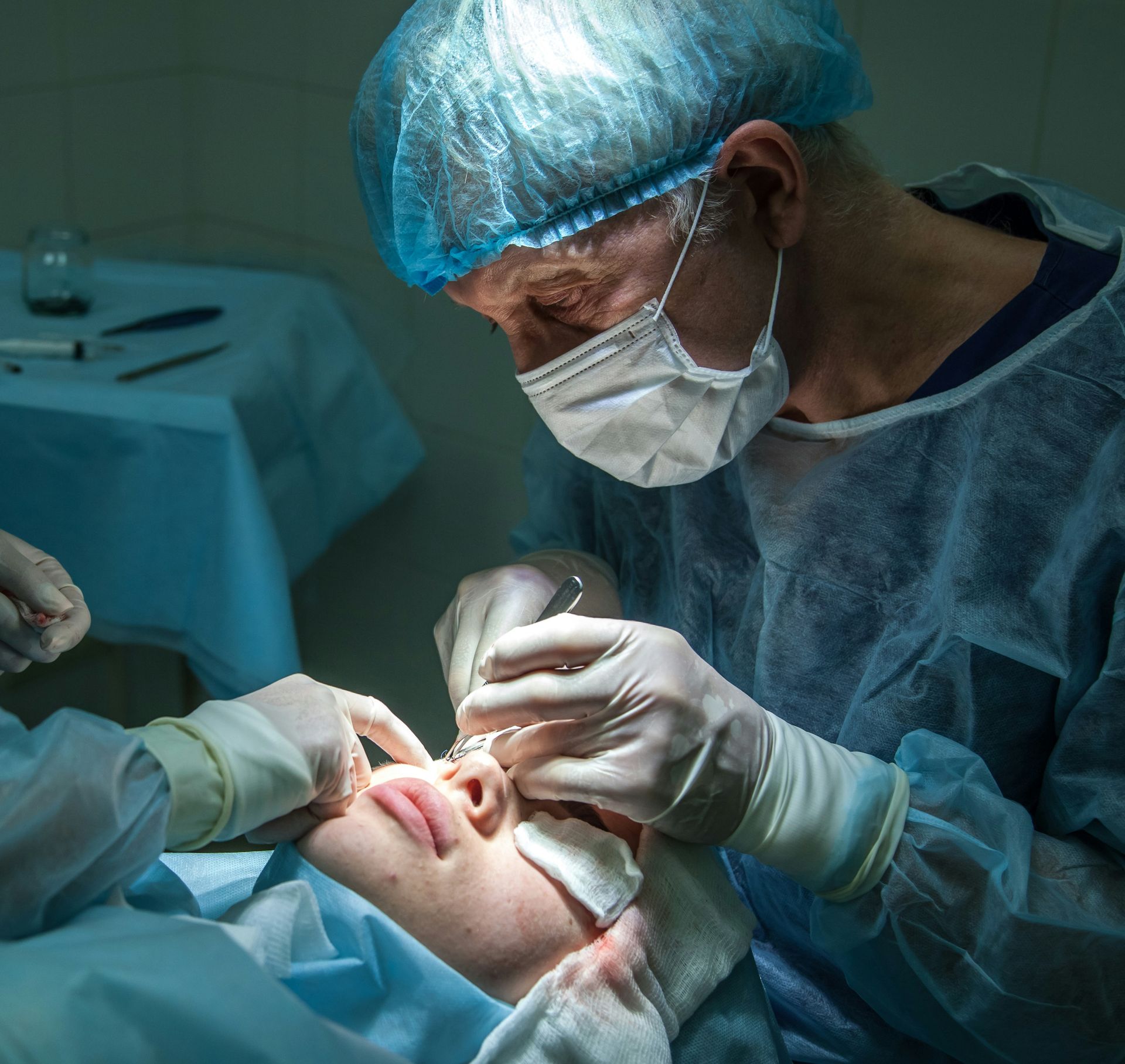 Un cirujano con bata azul y mascarilla realiza un procedimiento facial a un paciente en un quirófano.