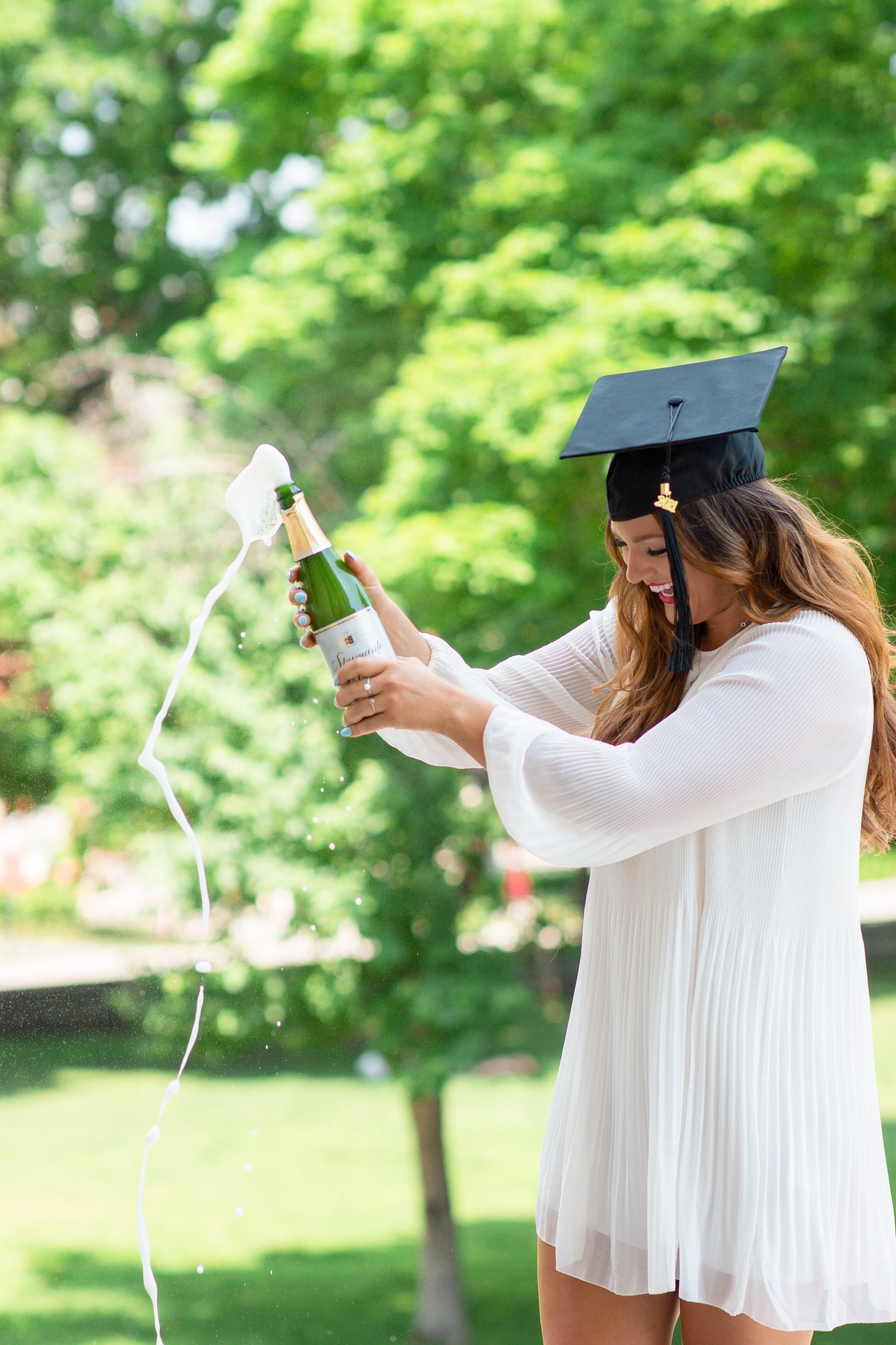 Graduada vestida de blanco rociando champán al aire libre en un parque verde.