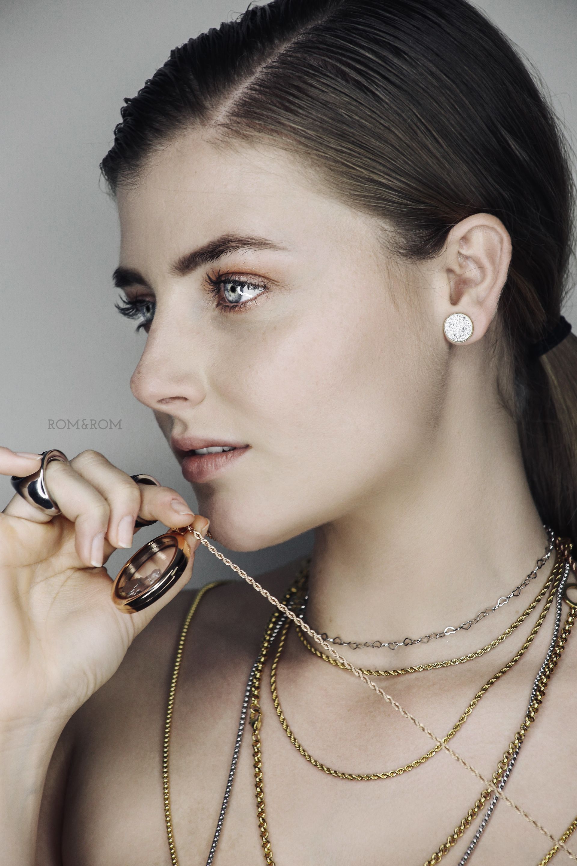 Perfil de una mujer con collares y pendientes de oro superpuestos, sosteniendo un frasco de perfume sobre un fondo oscuro.