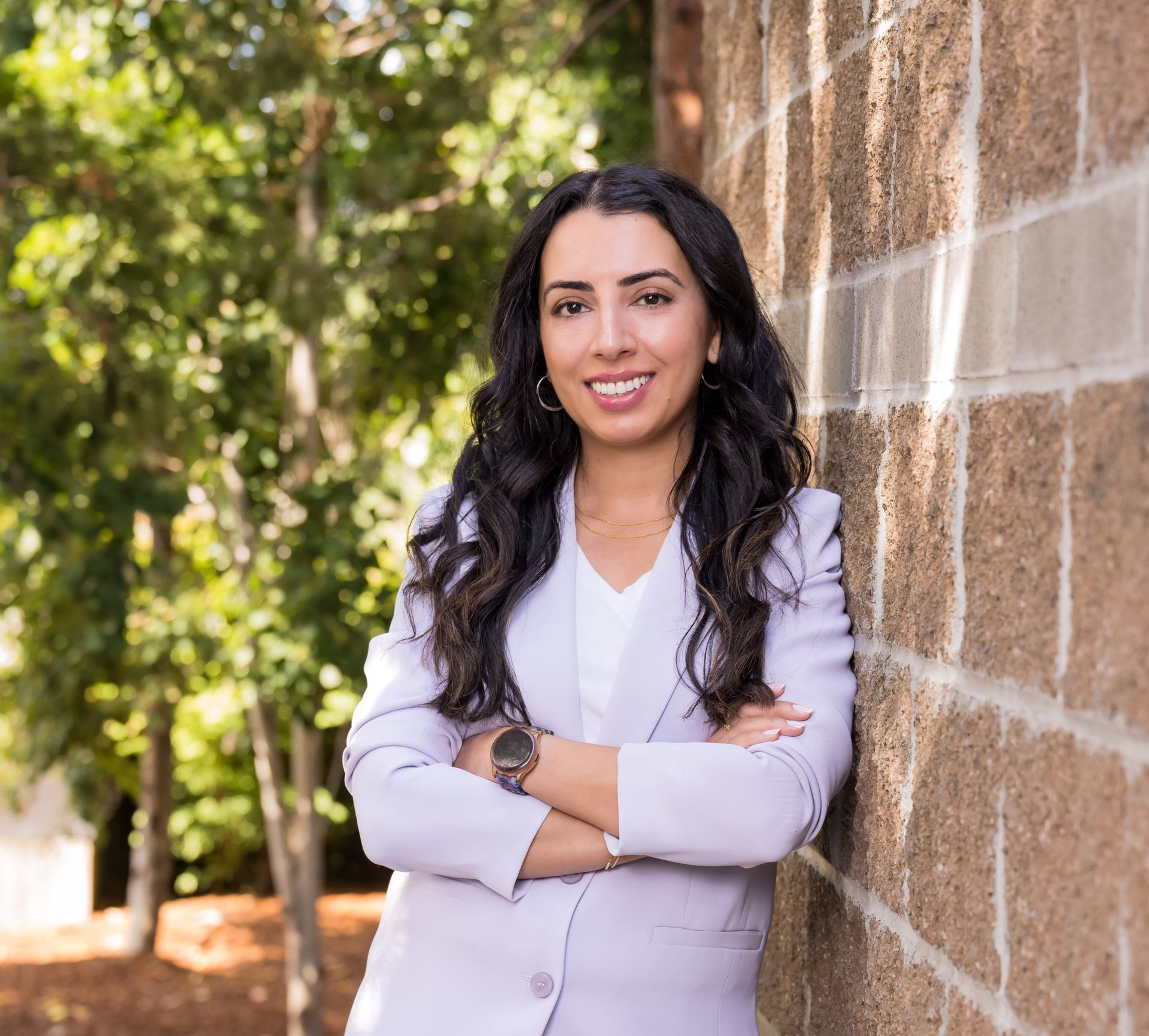 Anjabeen is leaning against a brick wall with her arms crossed.