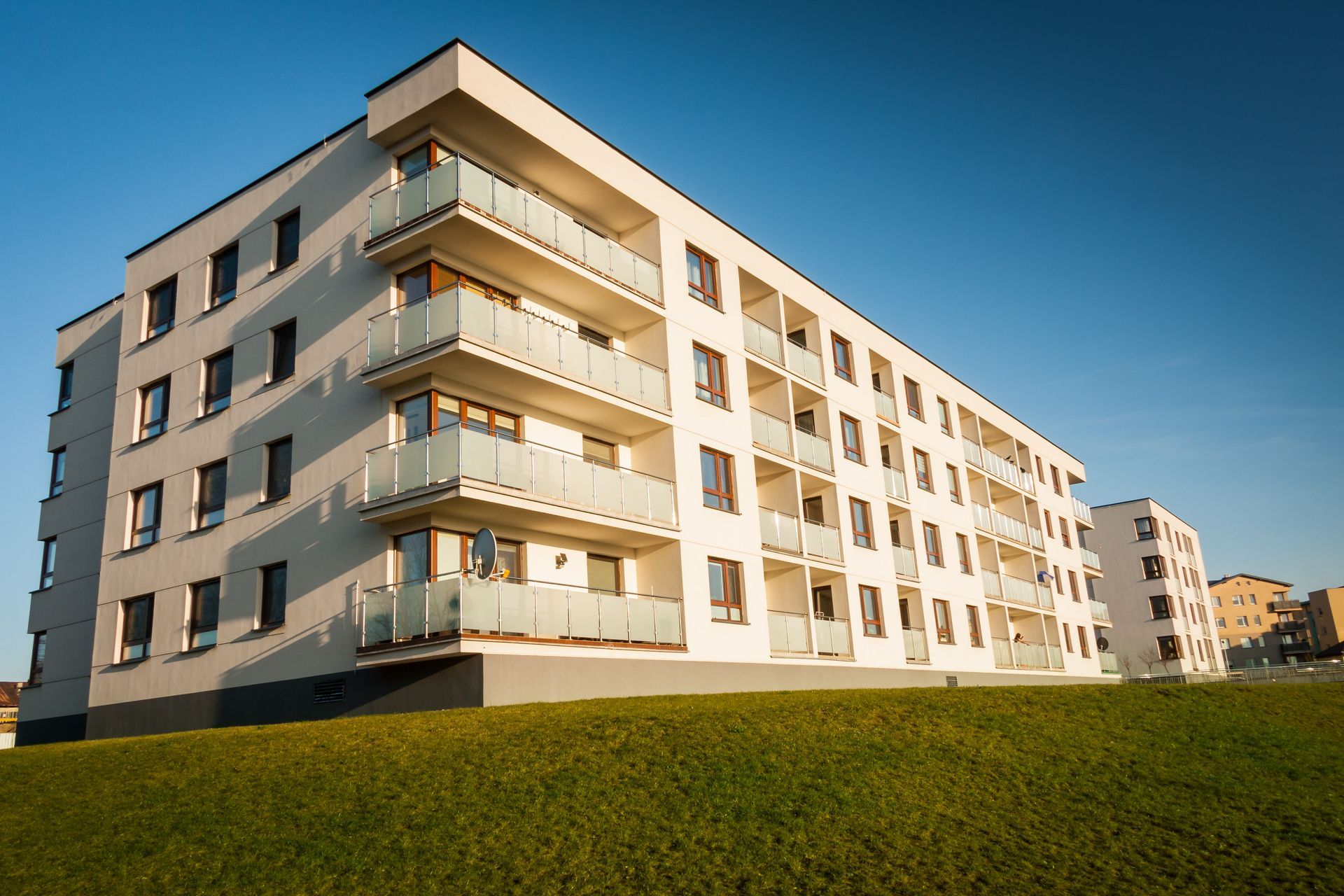 Un immeuble d'appartements moderne et blanc avec des balcons sous un ciel bleu vif, sur une colline herbeuse.