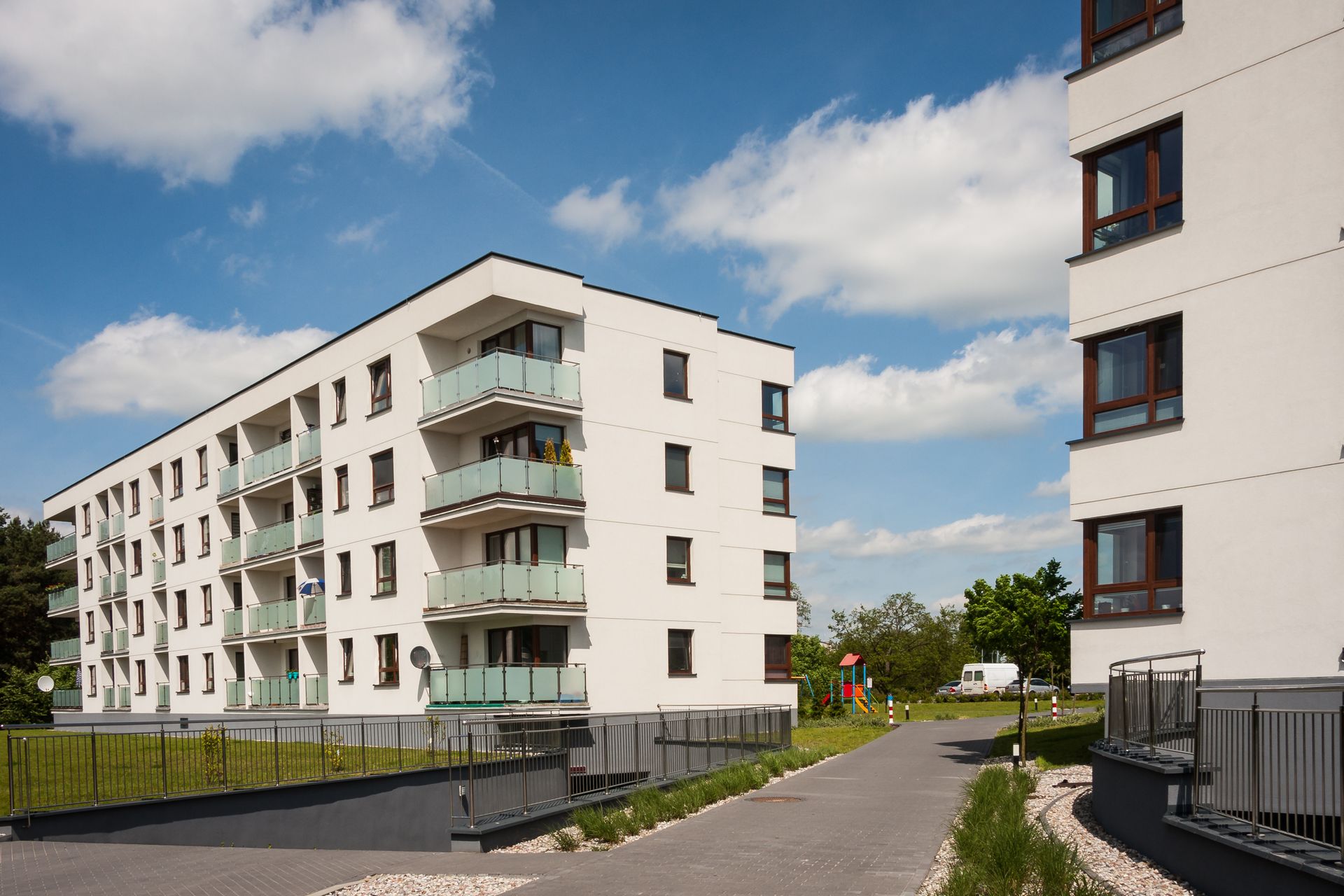 Des immeubles blancs avec balcons sous un ciel bleu parsemé de nuages. Un chemin pavé mène aux immeubles.