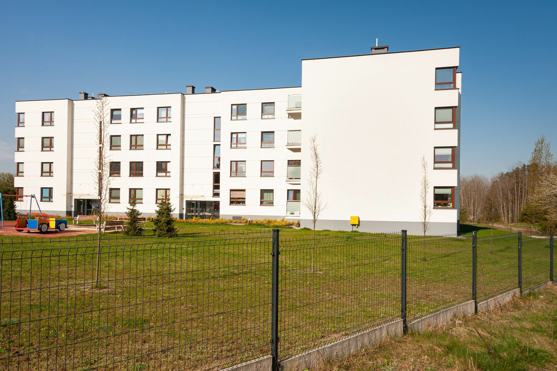 Immeuble blanc avec de nombreuses fenêtres, une pelouse verte, une aire de jeux et une clôture. Ciel bleu en arrière-plan.