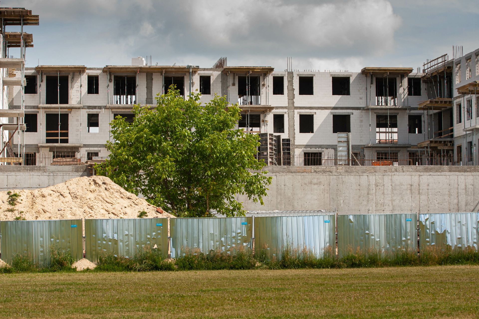 Bâtiment en construction avec des murs blancs inachevés, un arbre vert et une clôture patinée.