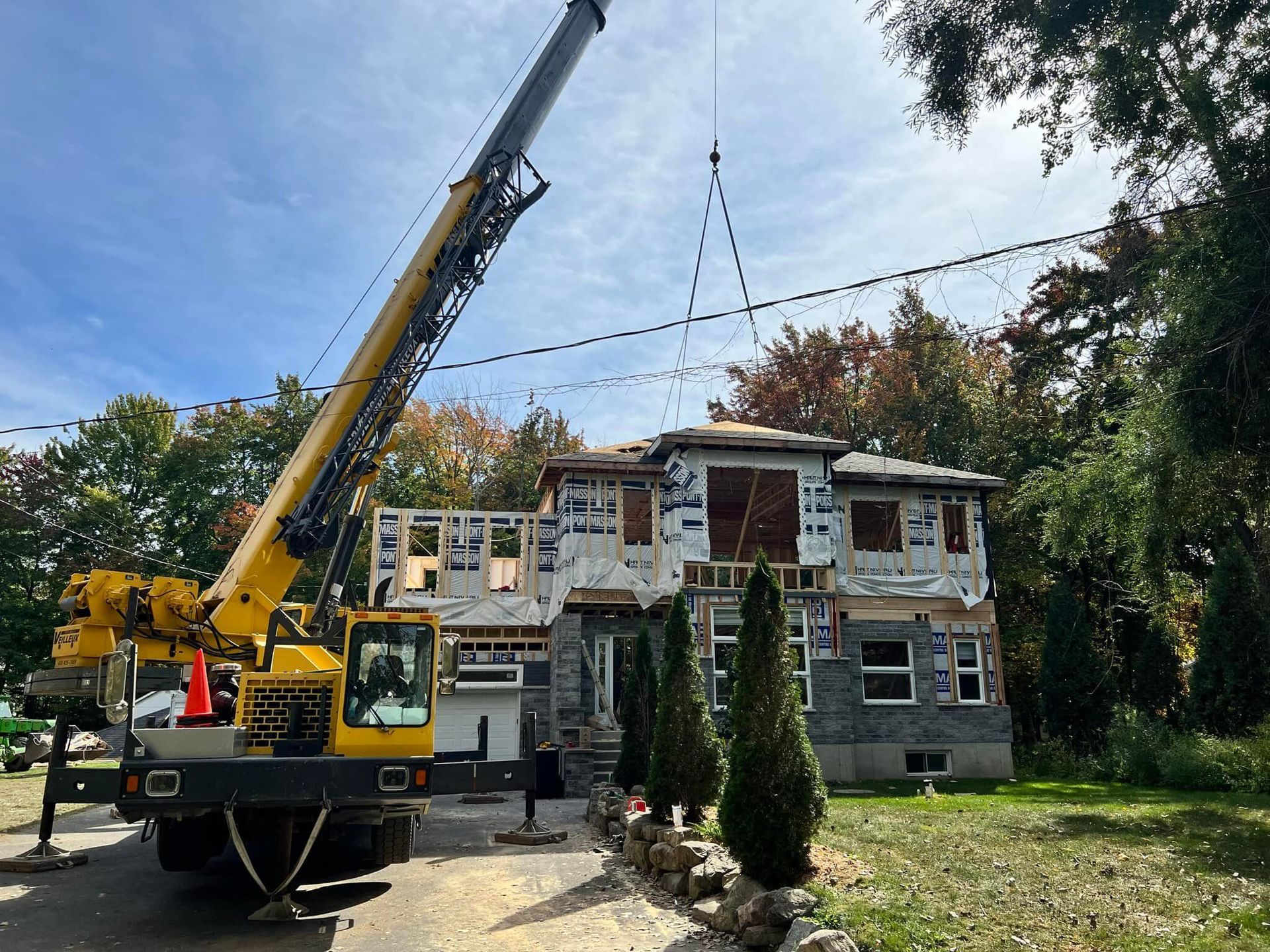 Une grue soulève une section de toit au-dessus d'une maison en construction. La charpente est apparente et certains murs extérieurs sont déjà terminés.