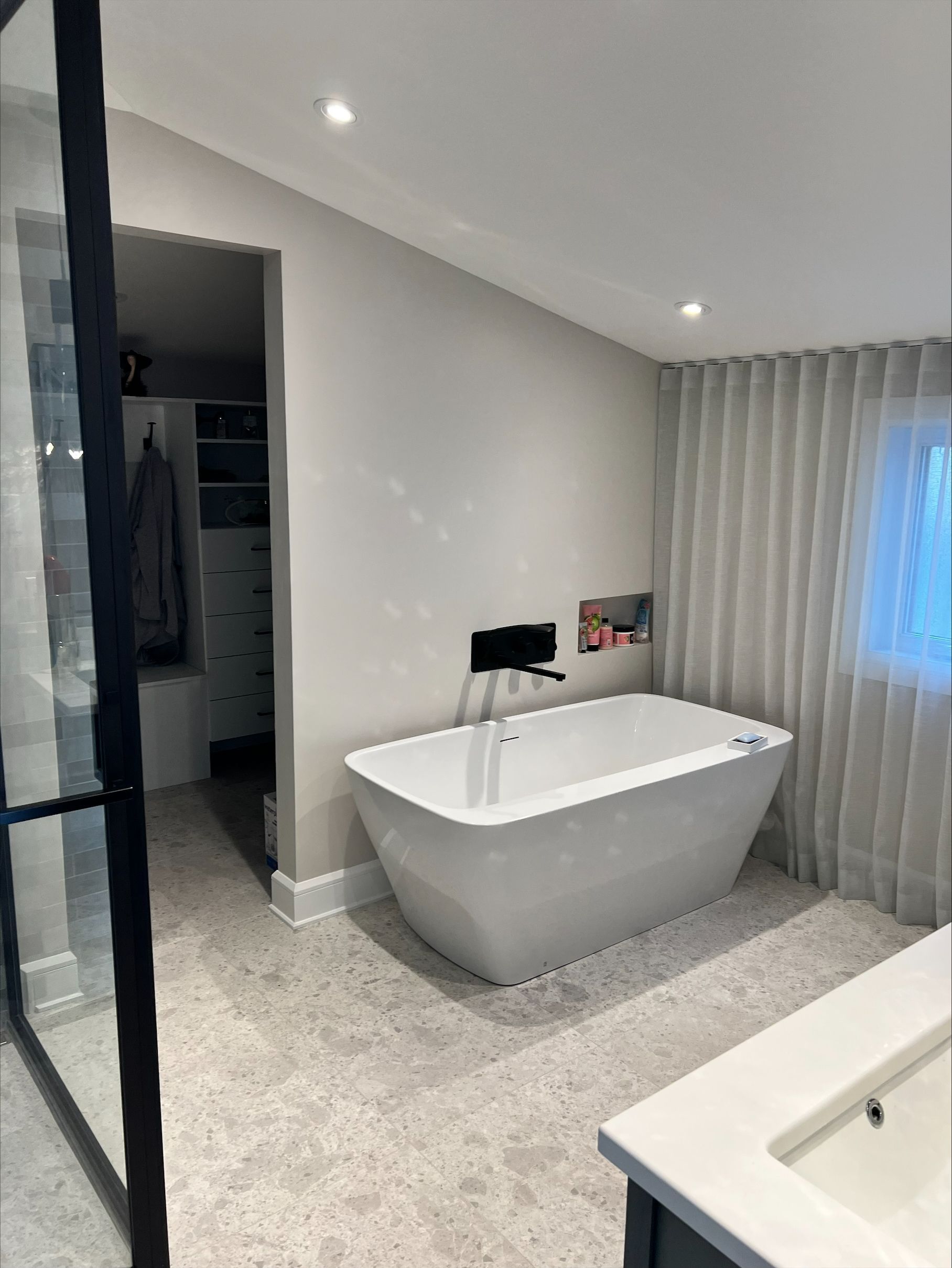 Salle de bain moderne avec une baignoire blanche autoportante, des murs neutres, un sol en terrazzo et un aperçu d'un dressing.