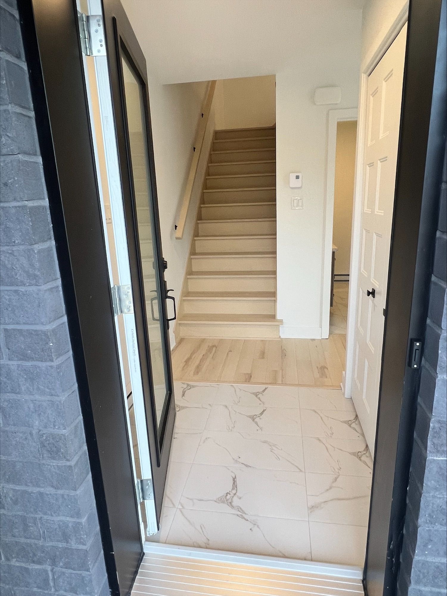Vue depuis une porte d'entrée ouverte sur un couloir avec un escalier menant à l'étage. Les sols sont carrelés et en bois.