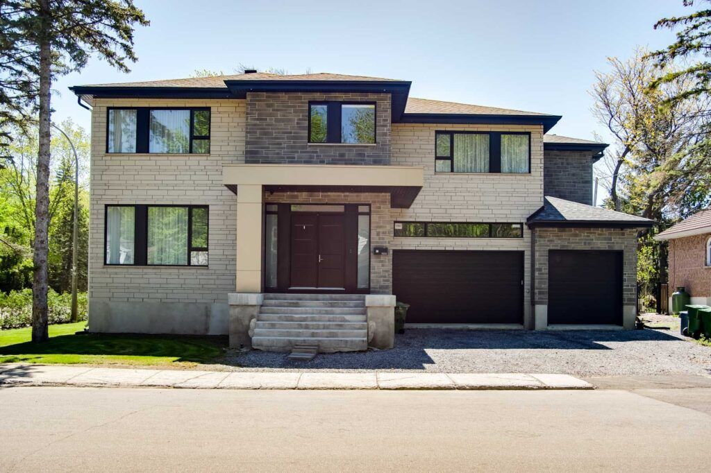 Maison moderne à deux étages avec une façade grise, des portes de garage marron foncé et une entrée principale avec escalier par une journée ensoleillée.