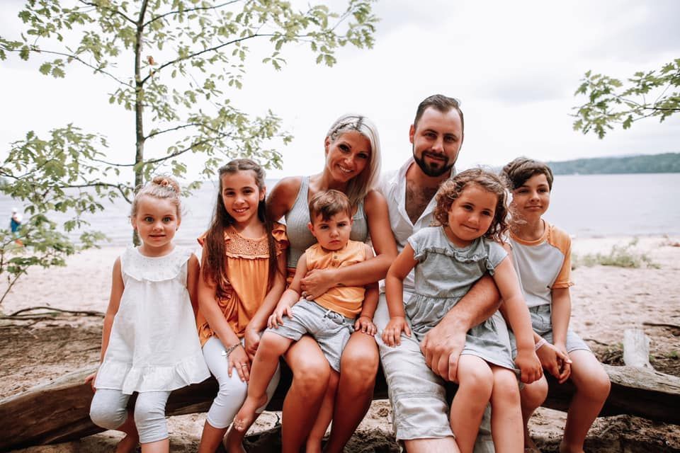 Une famille de six personnes souriant et posant dehors, près d'un lac. Les parents et les enfants sont assis sur un banc rustique sous les arbres.