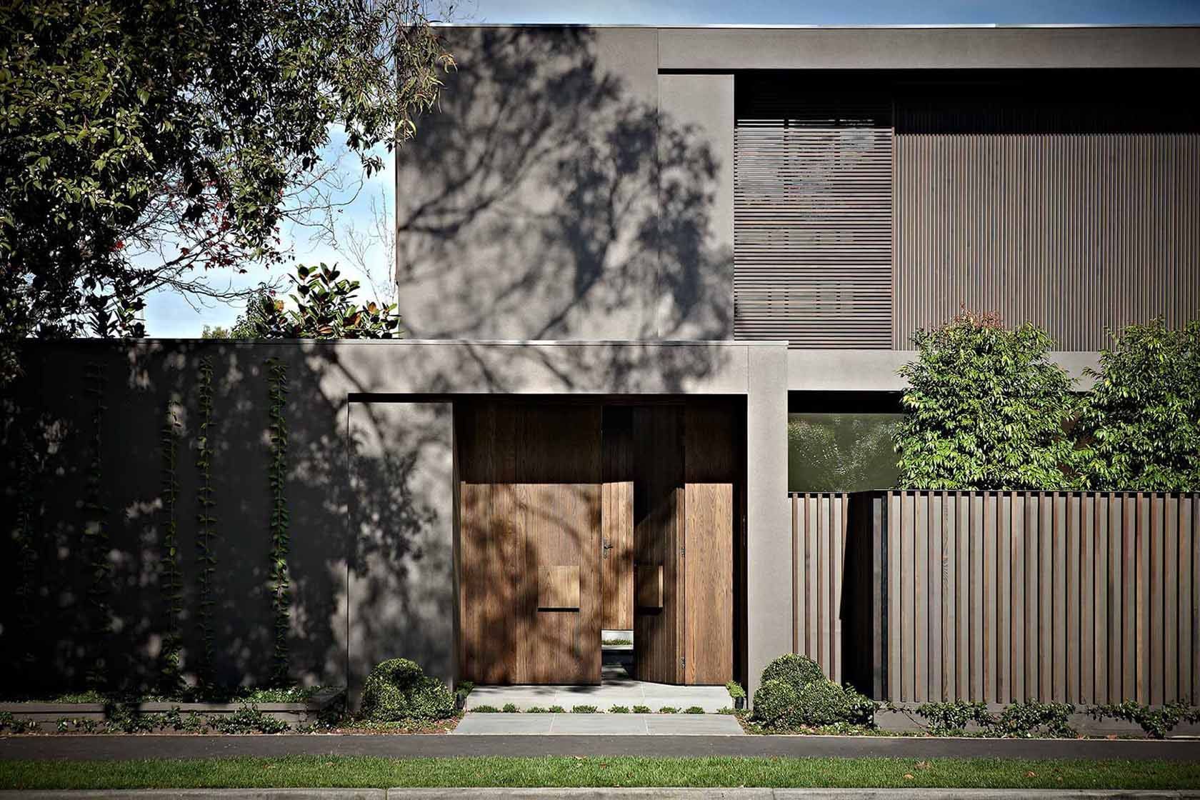 Extérieur d'une maison moderne avec une porte en bois, des lattes de bois verticales et une façade en béton avec des ombres d'arbres.