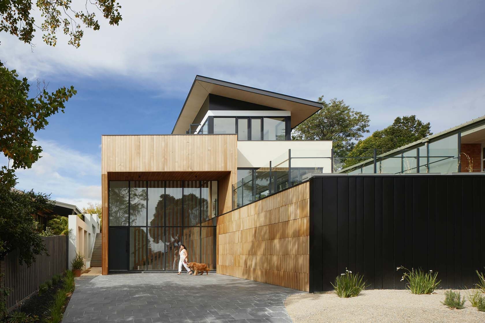 Maison moderne à deux étages avec façade en bois et noire. Une personne avec un chien se tient près de l'entrée vitrée par une journée ensoleillée.
