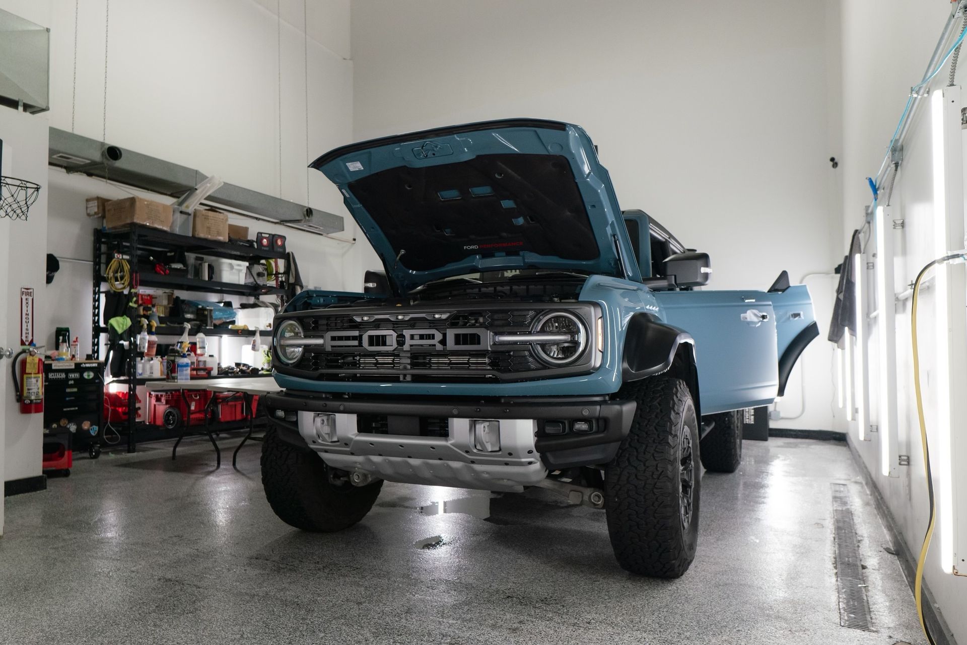 A blue ford bronco is parked in a garage with its hood up.