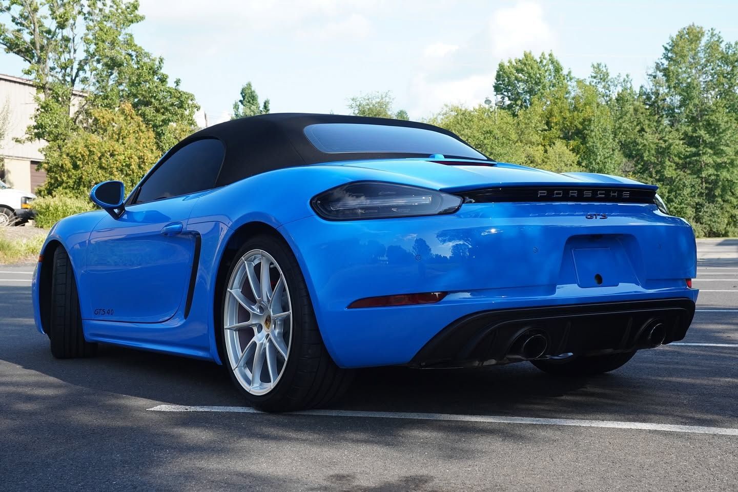 Blue convertible sports car parked on a road beside trees, viewed from the rear side.