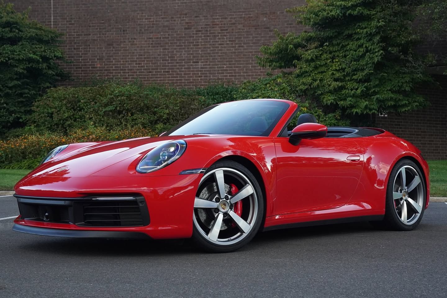 Red convertible sports car parked on pavement in front of a brick wall and greenery
