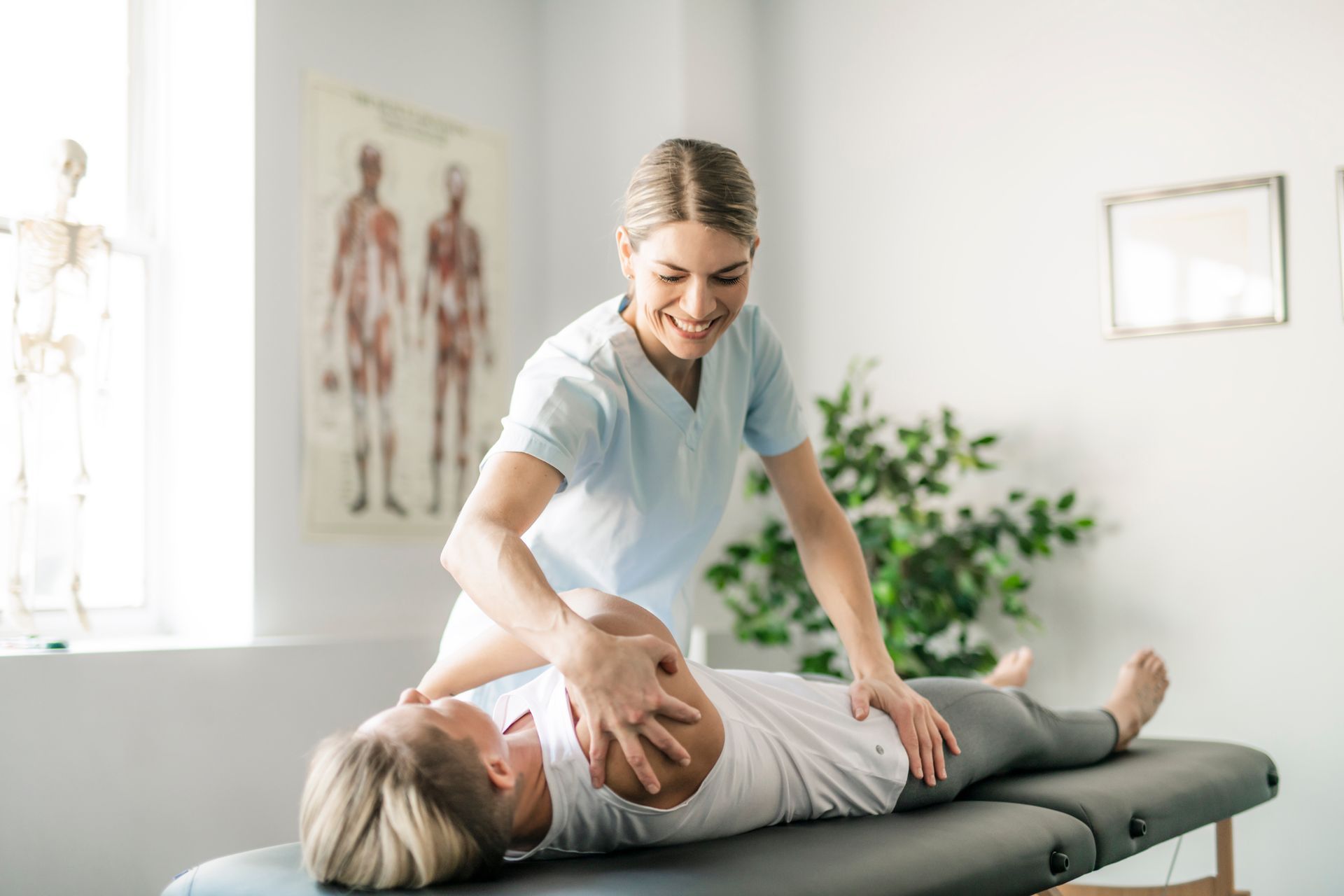 Physical therapist assisting a patient with arm movement on a treatment table. Smiling therapist, light-filled room.