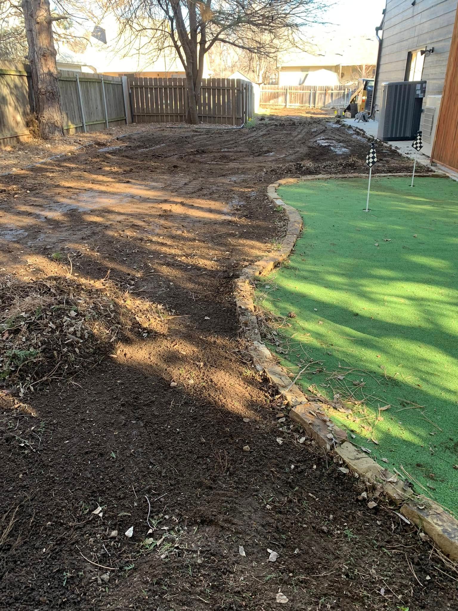 Muddy backyard with artificial turf border, tree, and wooden fence.