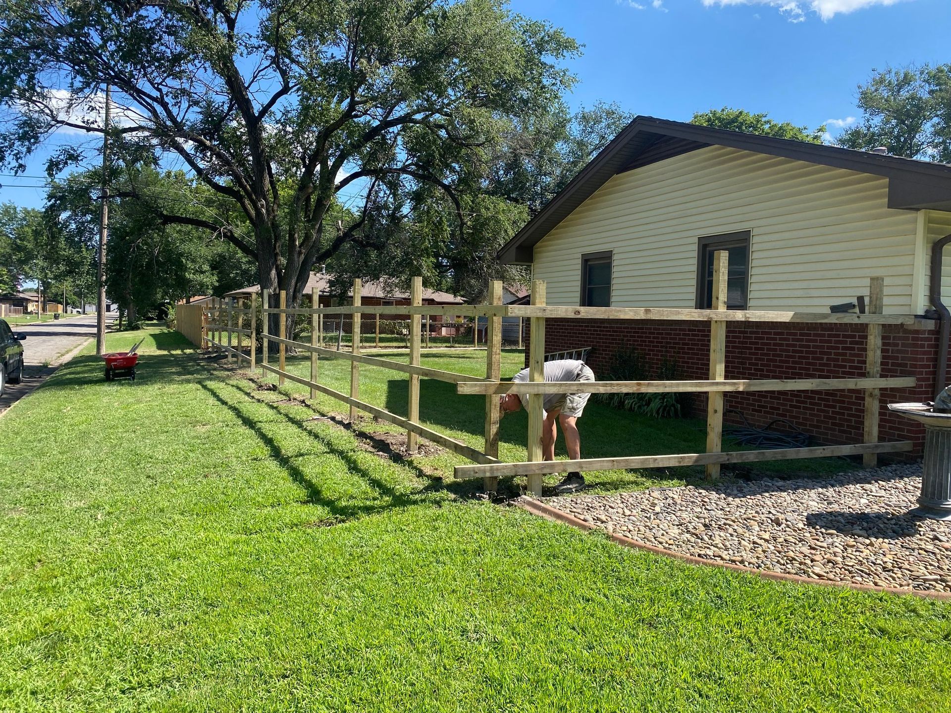 Person building a wooden fence in a yard with green grass. A tan house and street are in the background.