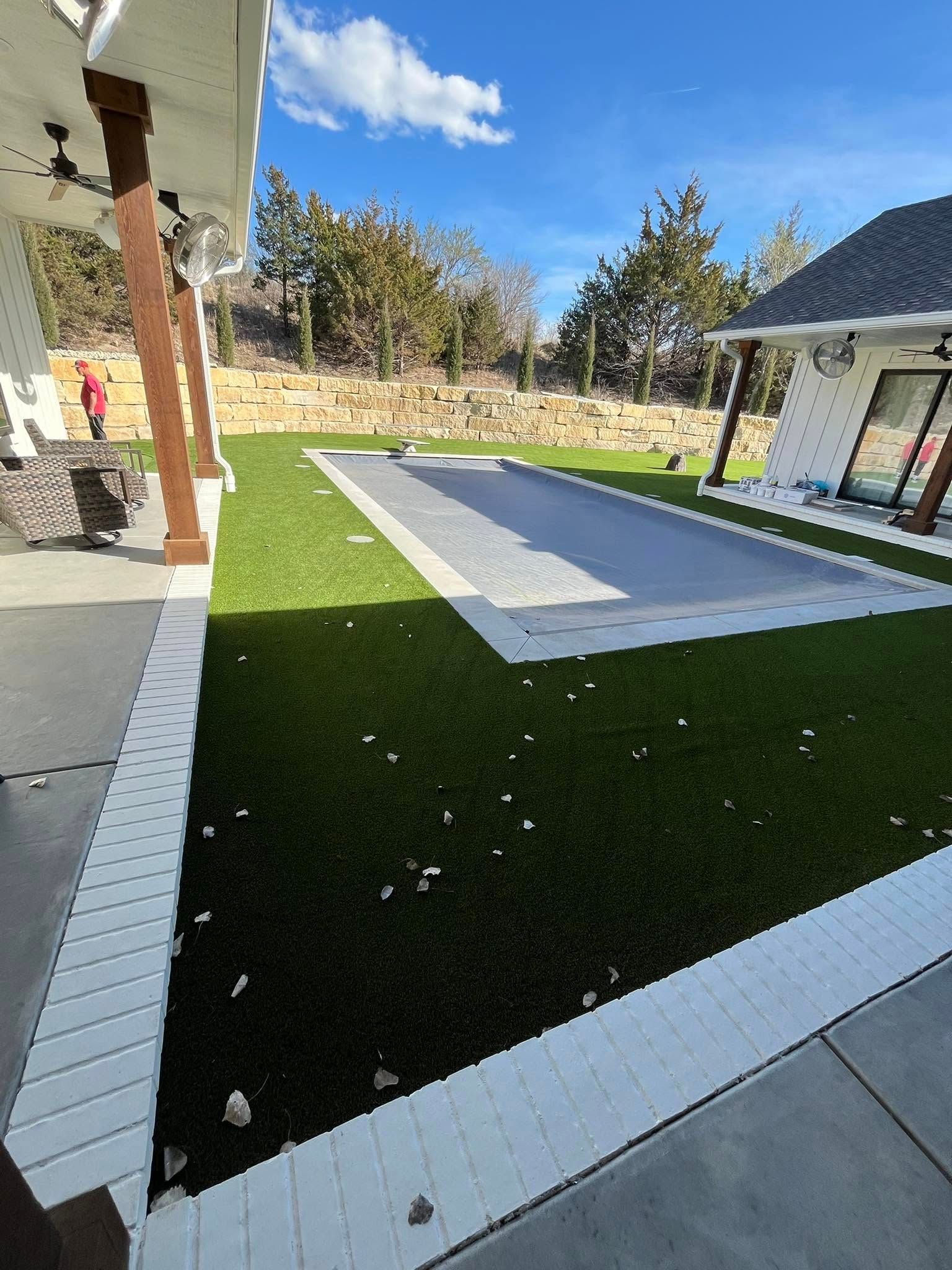 A backyard with artificial turf, a covered pool, and a house under a blue sky.
