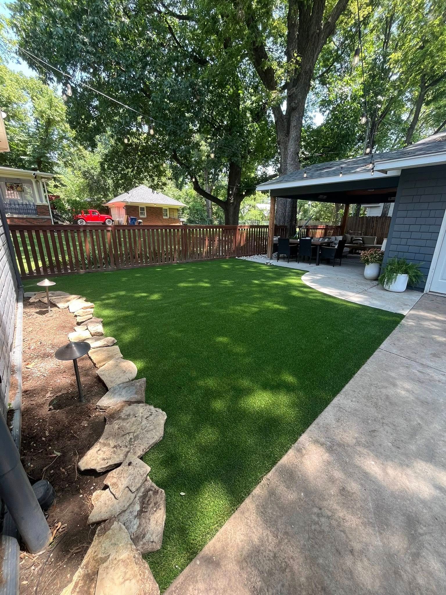 A backyard with green grass, stone landscaping, and a covered patio. Wooden fence and trees in the background.