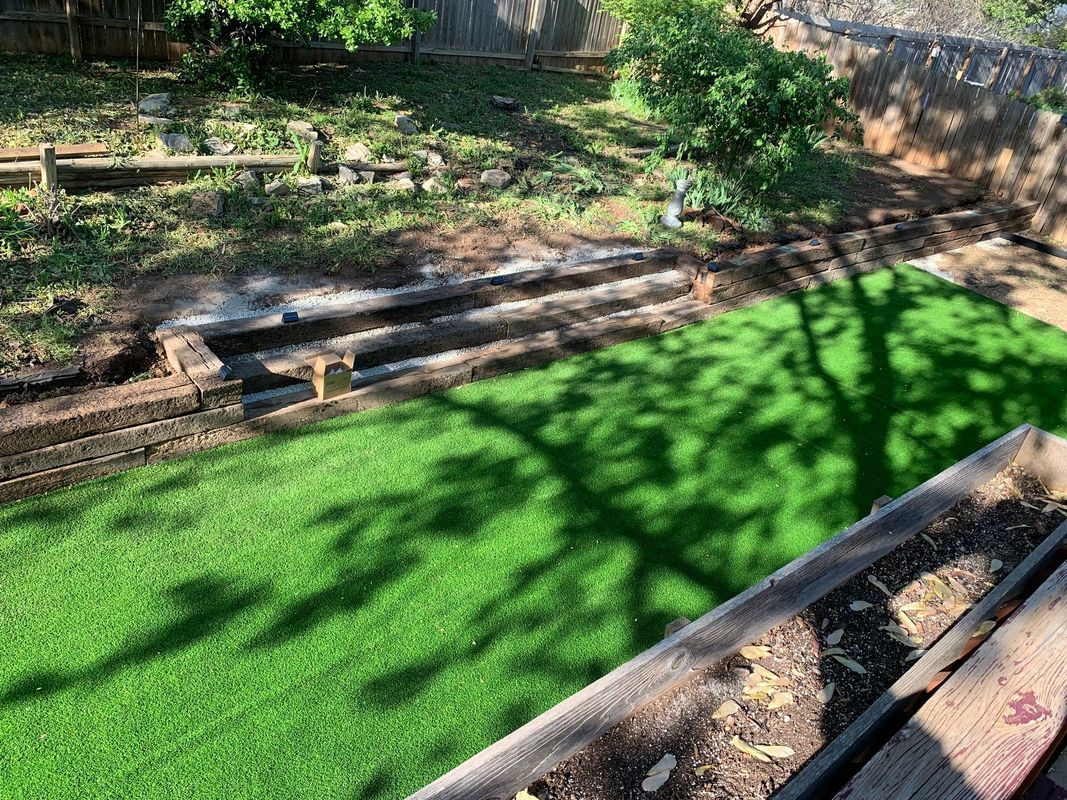 Backyard with new artificial turf, dirt areas, and wooden structures. Bright green lawn, sunny day, shadow.