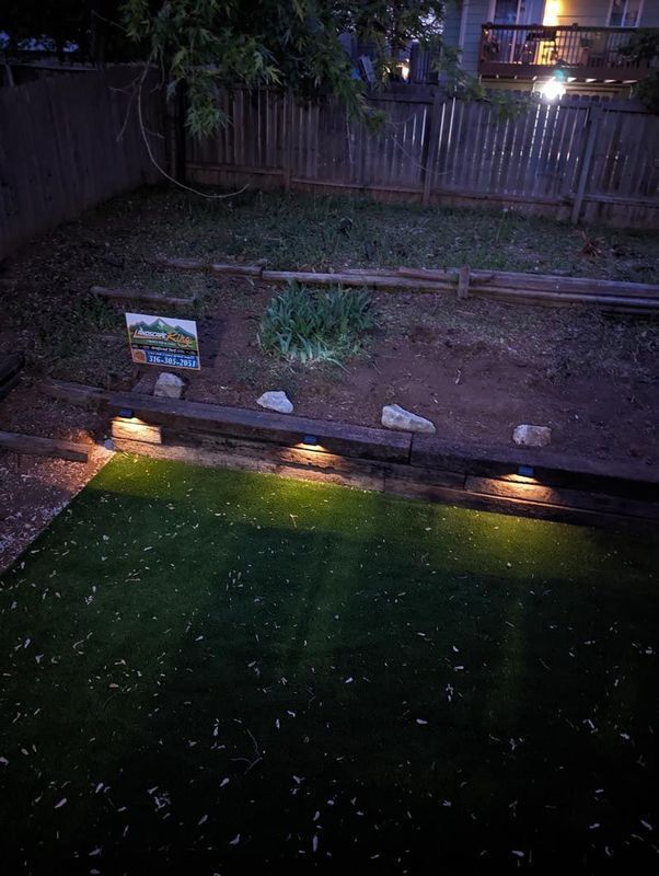Backyard at night, with artificial grass, lighting, a wooden fence, and a painting.