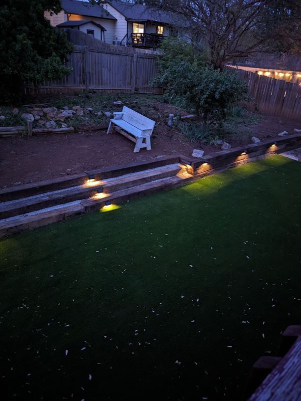 Backyard scene at dusk. White bench on a stone patio, illuminated steps and lawn. String lights.