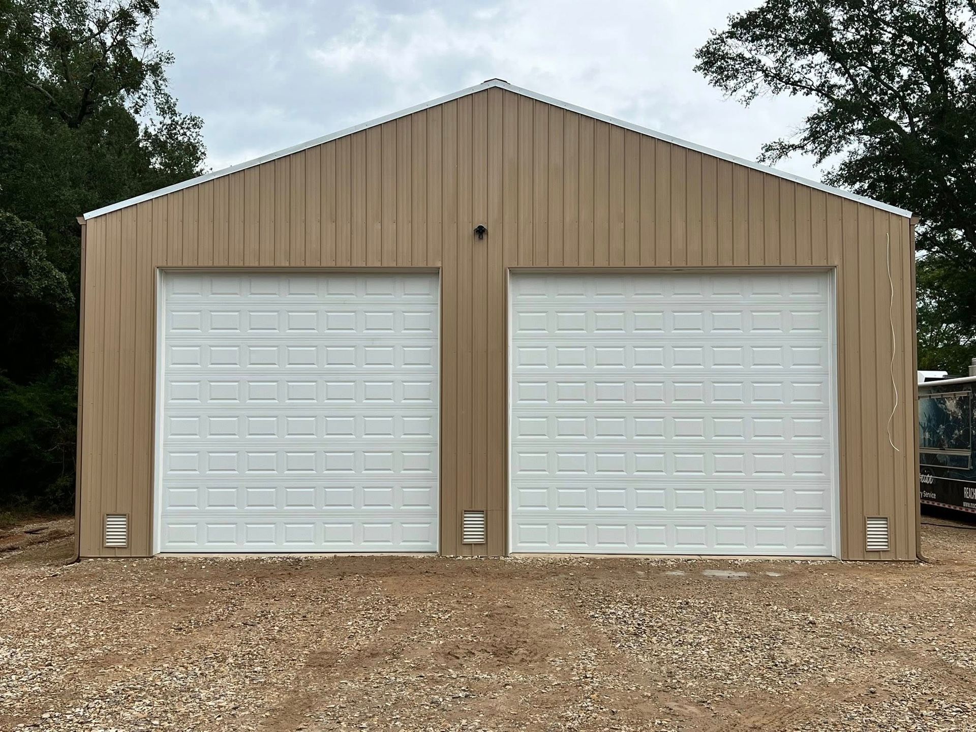Tan metal two-car garage with white garage doors, set on gravel.