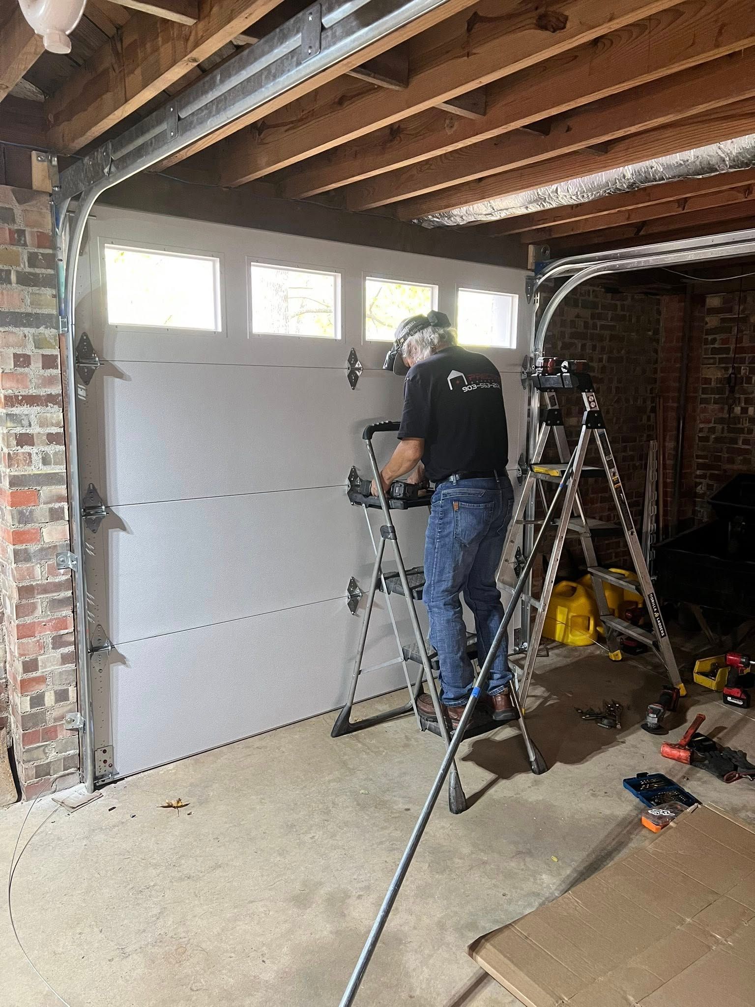 Person on a ladder installing a white garage door, indoors. Brick wall on the left.