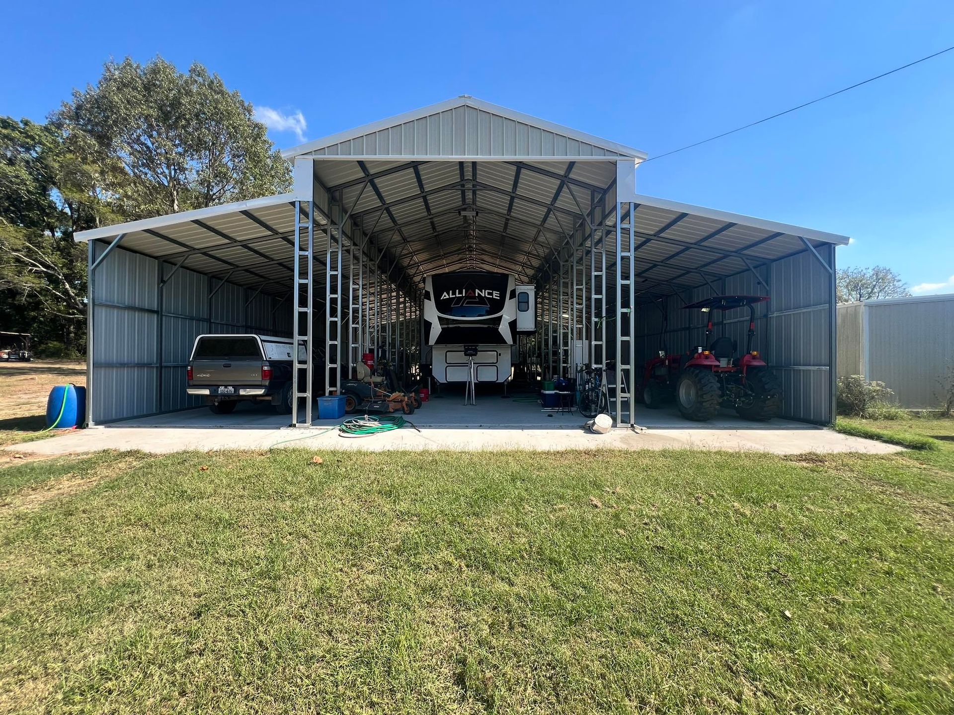 Metal carport shelter with vehicles, including an RV, truck, and tractor, parked on a concrete slab.