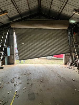 Garage interior with a roll-up door partially open. Concrete floor, metal roof, and sunlight from outside.