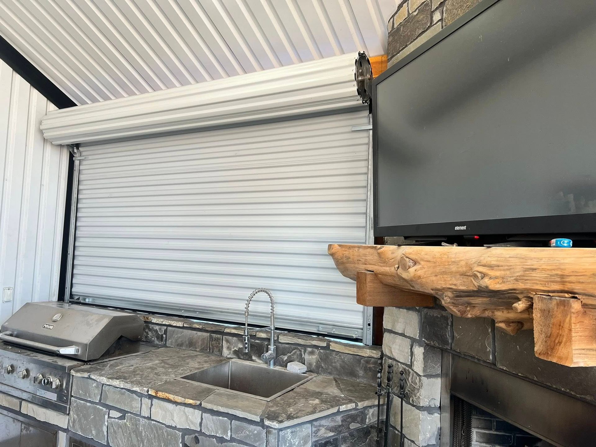 Outdoor kitchen with a roll-up shutter covering an opening, next to a built-in grill and sink.