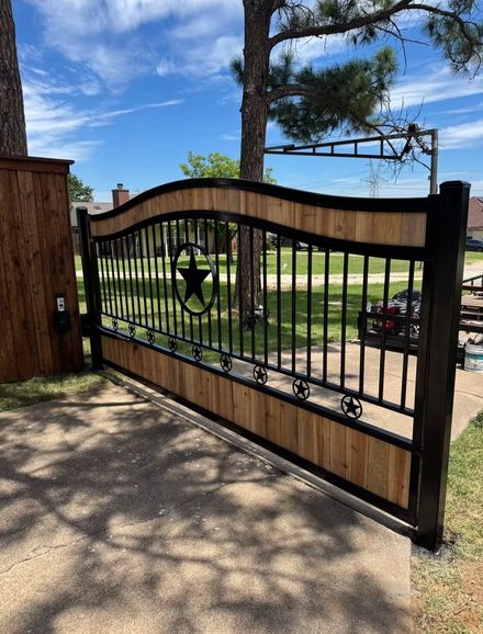 A wooden and black metal gate with a Texas star design is in front of a driveway, on a sunny day.
