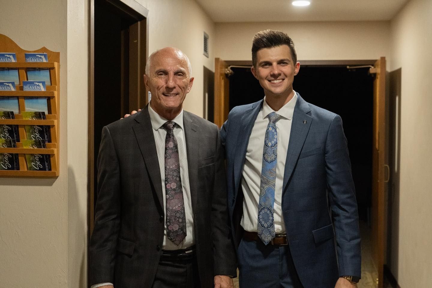 Two men in suits pose in a hallway. One man puts his arm around the other’s shoulders.