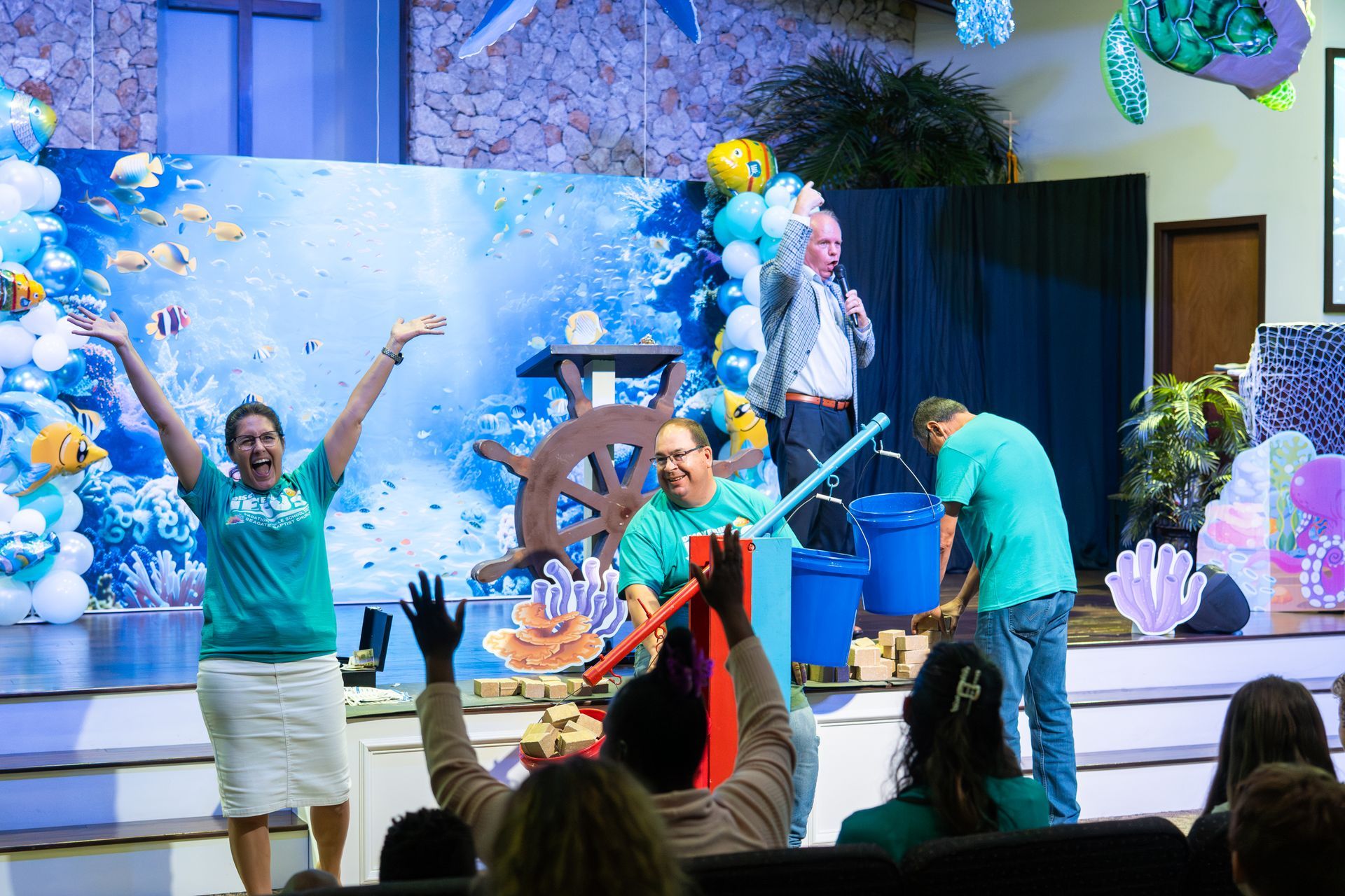 A man gestures on a stage with castle backdrop, knights, and an audience of children.