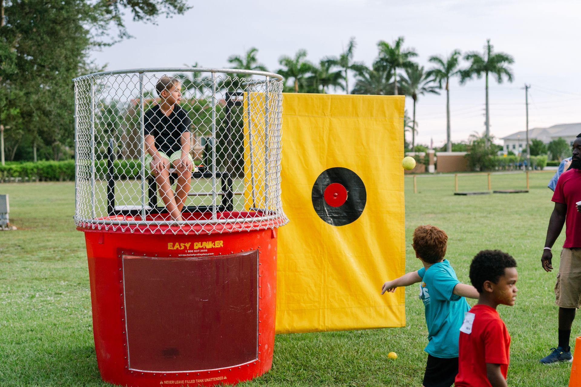 Boy about to throw a ball at a dunk tank, yellow target in background; outdoors on grass.
