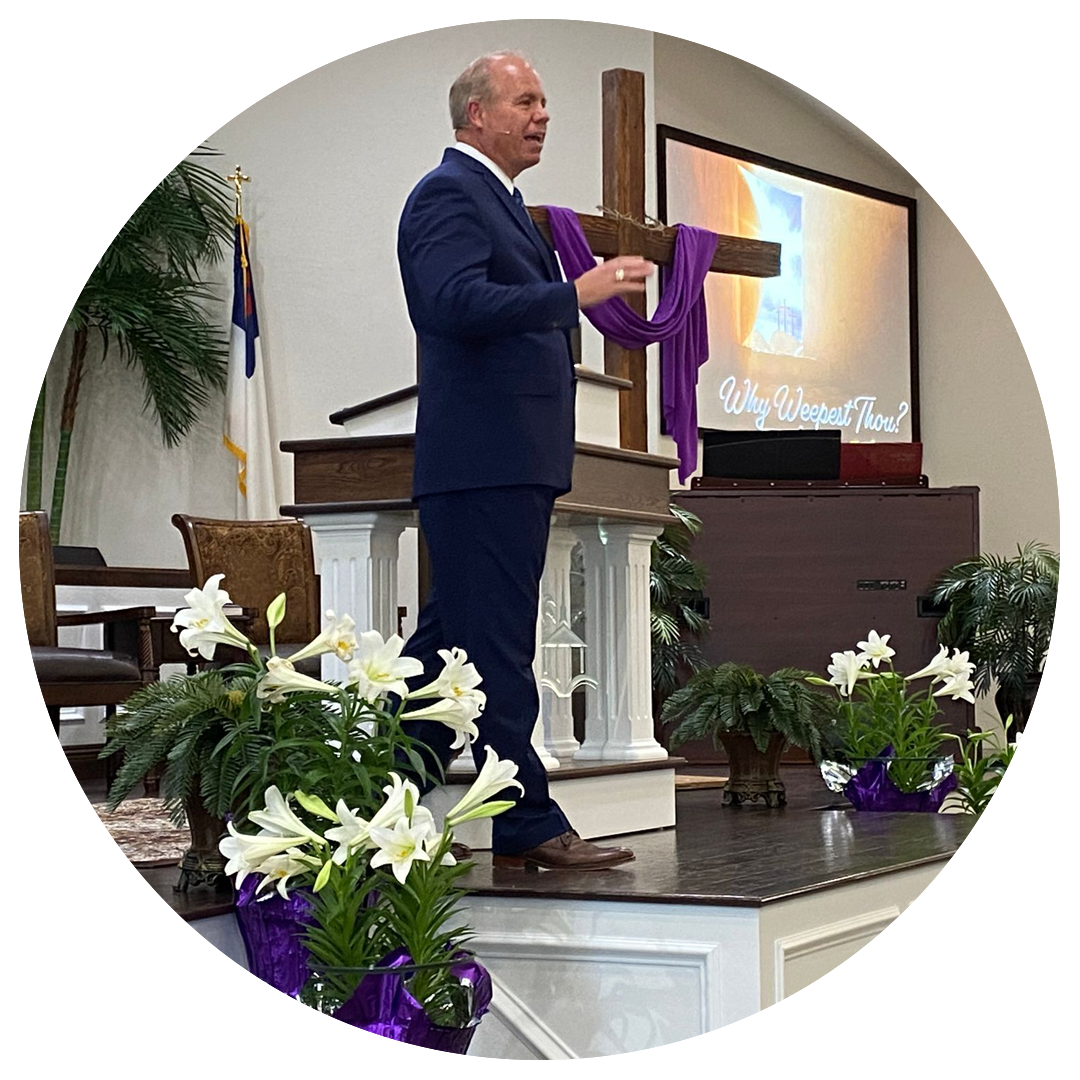 Man in suit preaching at a church, cross on the stage, lilies in front.