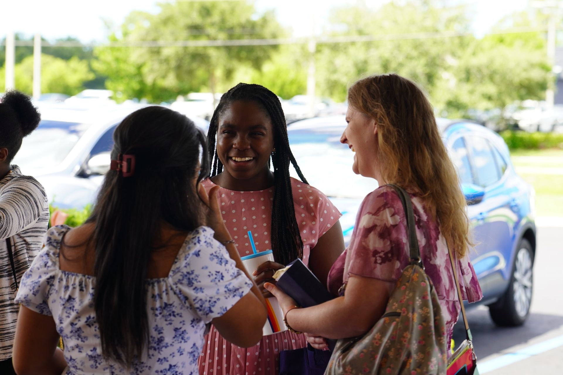 Three smiling women outside, greeting each other. One woman has long braids, holding a drink and a book.