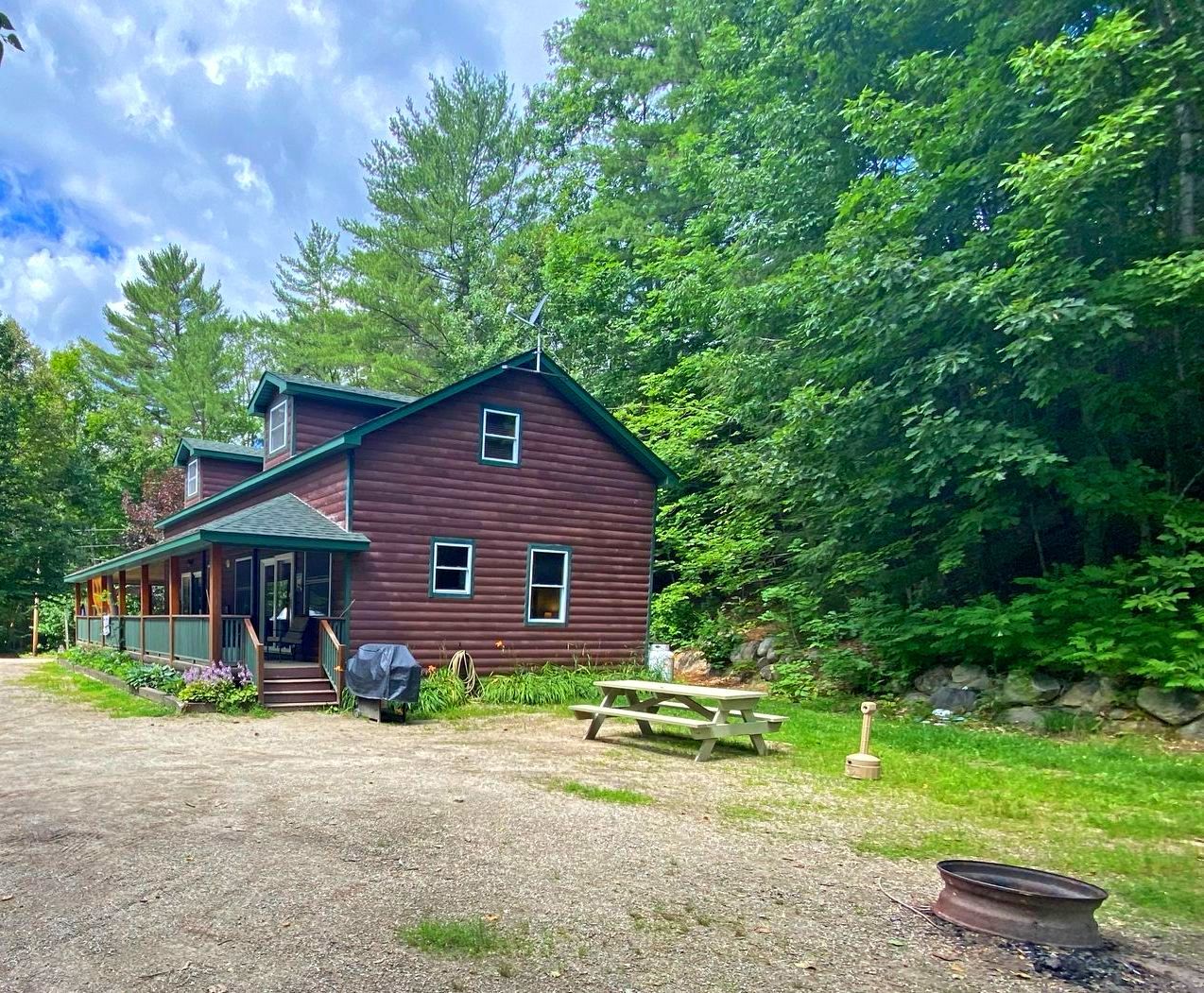 A large log cabin in the middle of a forest with a picnic table in front of it.