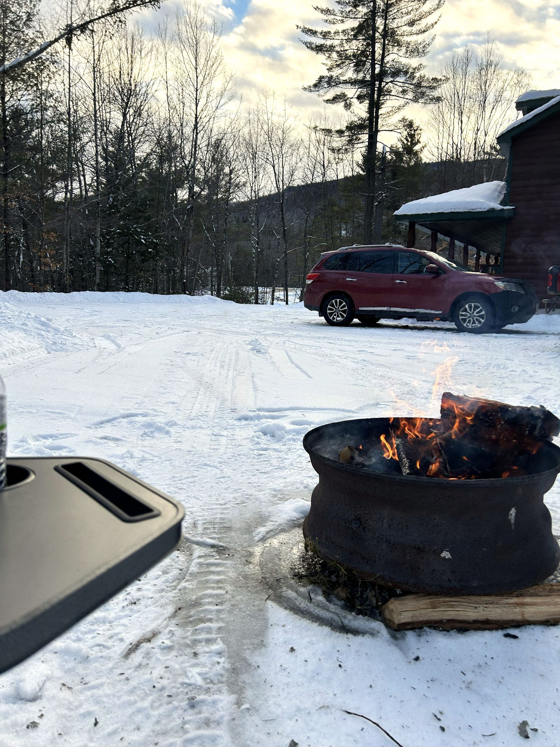 Snowy driveway with a red SUV, a fire pit burning in the foreground, and trees in the background