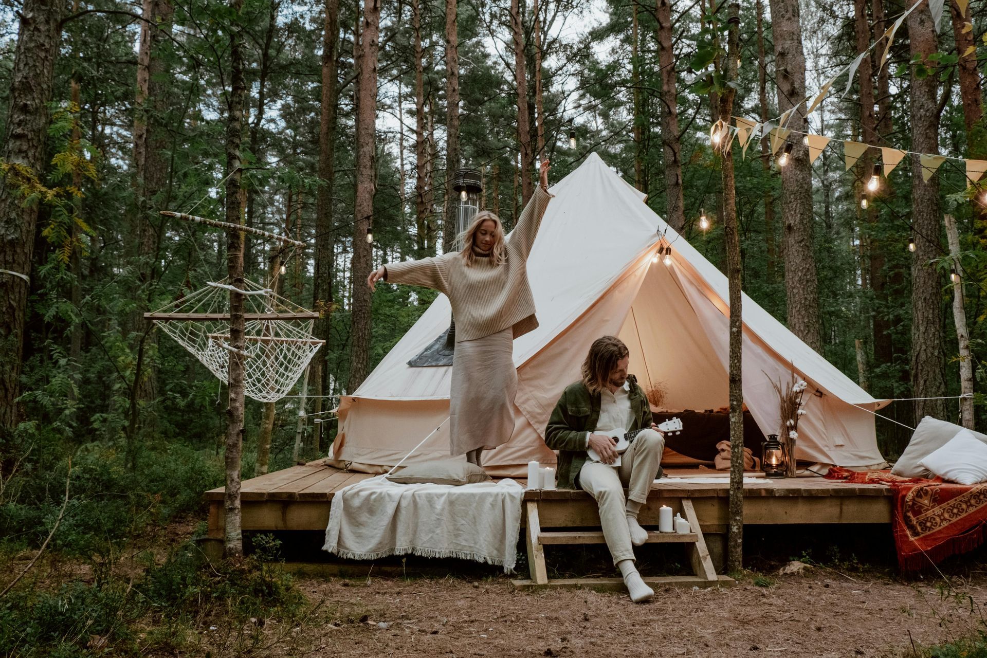 A man is playing a guitar while a woman stands in front of a tent in the woods.