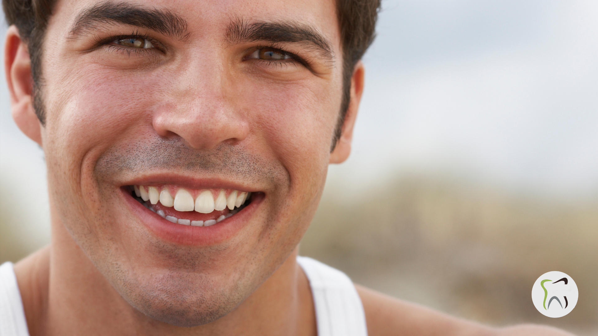 Man smiling broadly, white teeth visible, wearing a white tank top, outdoors.