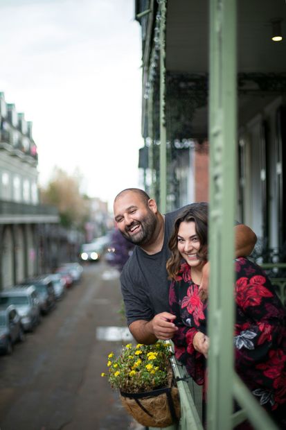 Couple smiling on a balcony overlooking a city street. Balcony has hanging flowers.