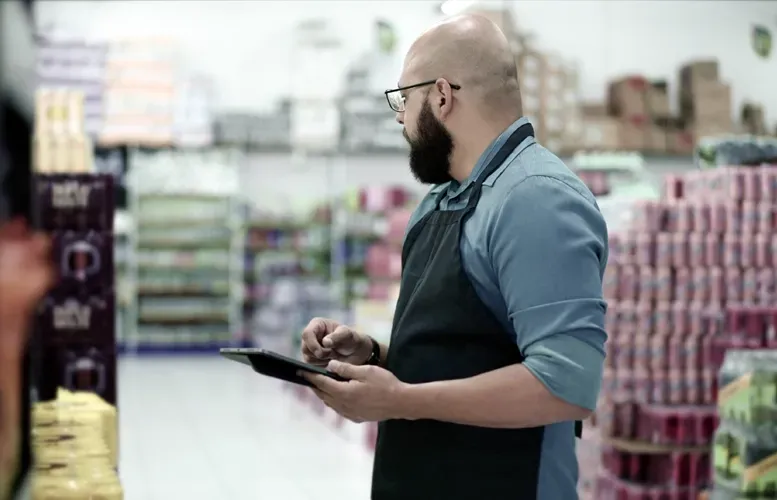 Man in apron, holding a tablet, checking inventory in a grocery store.